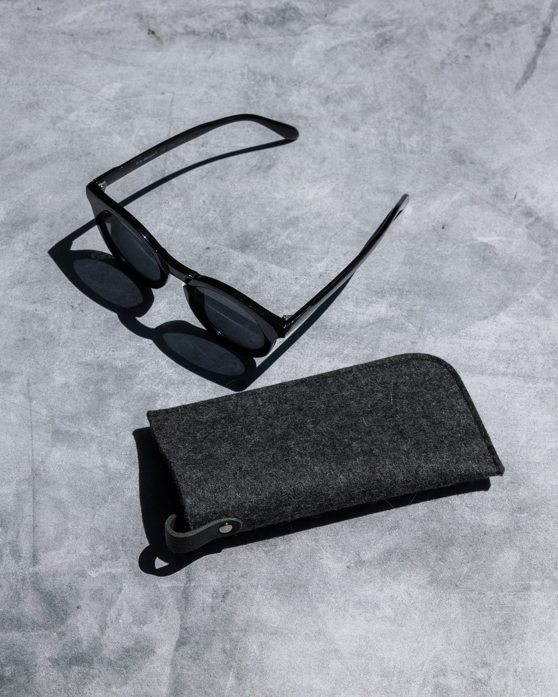 A pair of black sunglasses sits on a gray concrete surface, alongside a graf lantz Merino wool eyeglass sleeve for optimal protection.