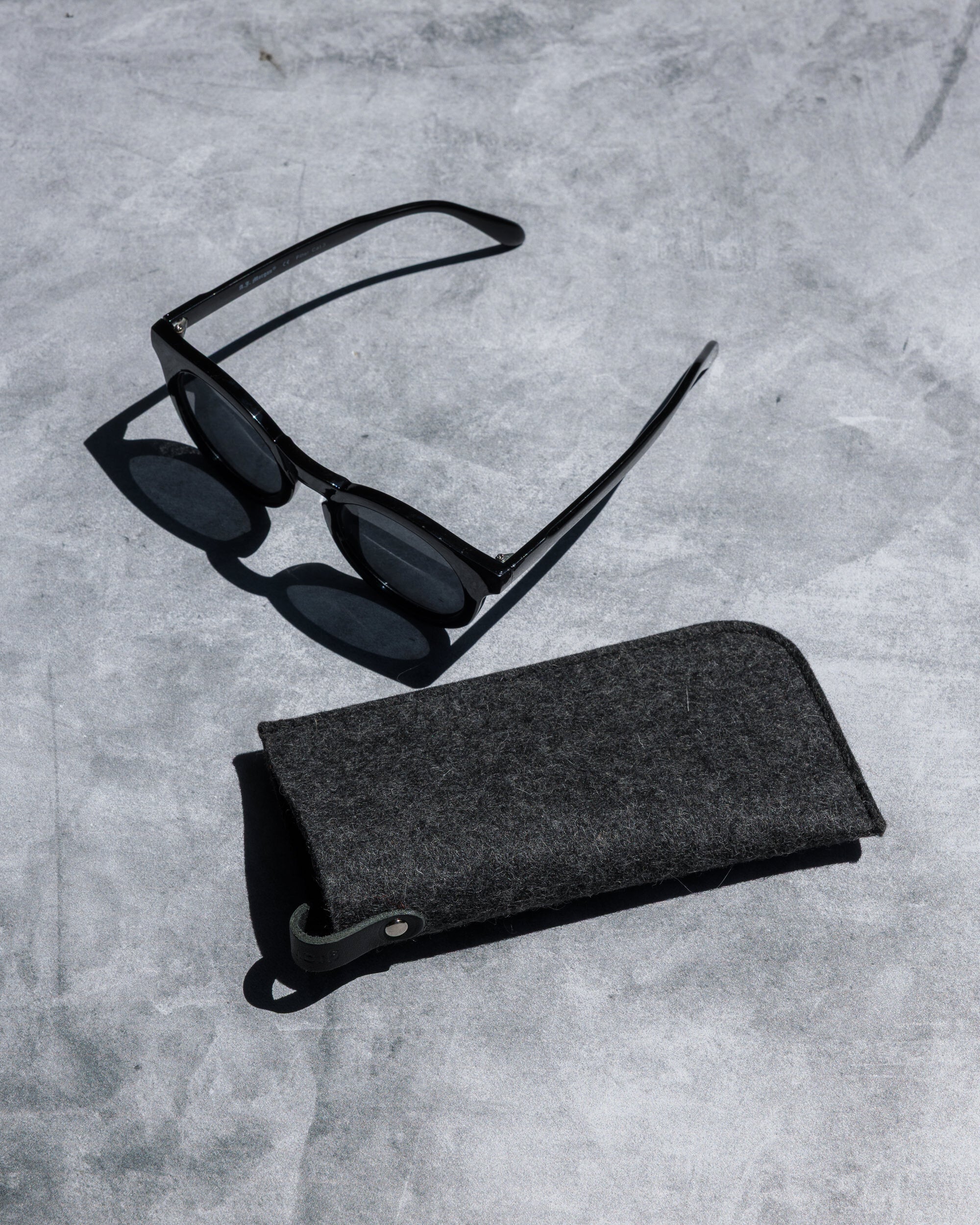A pair of black sunglasses sits on a gray concrete surface, alongside a graf lantz Merino wool eyeglass sleeve for optimal protection.