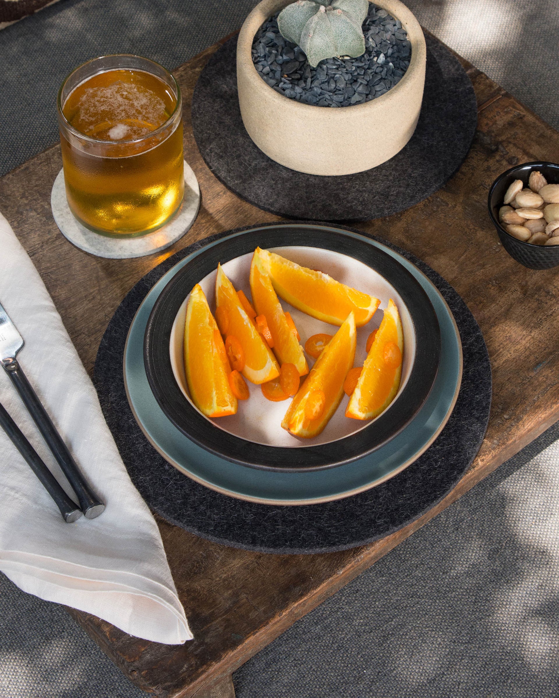 A wooden table holds a plate of orange slices, a glass of beer, almonds in a bowl, and a beige pot with a succulent. Nearby are a knife, fork, and moisture-wicking merino wool cloth napkin by graf lantz. Shadows dance across the heat-resistant surface from adjacent foliage.