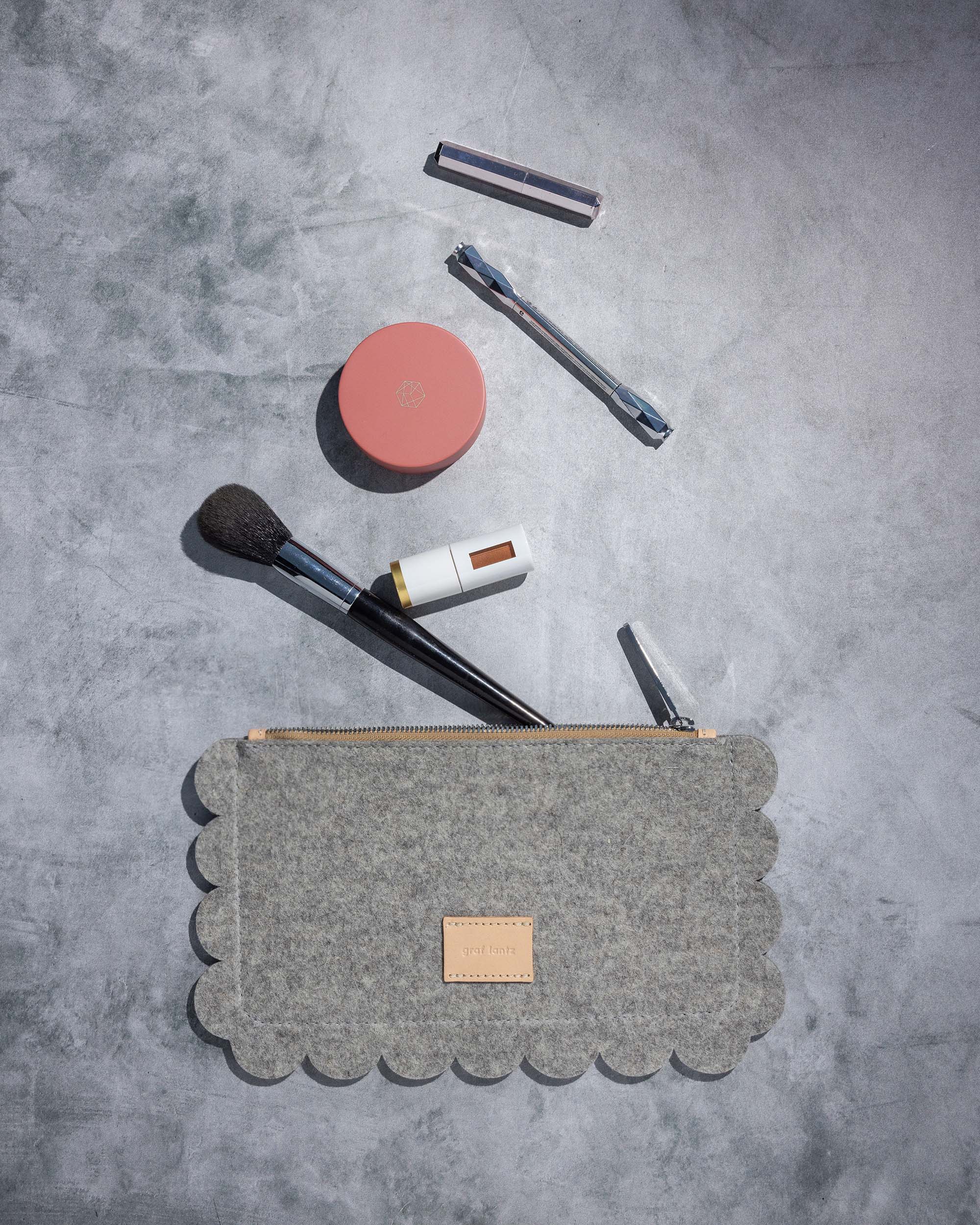 A flatlay features an open graf lantz Deco Edge pouch, a round blush compact, a makeup brush, and two lipstick tubes on a textured gray surface. The neat arrangement highlights visible shadows.