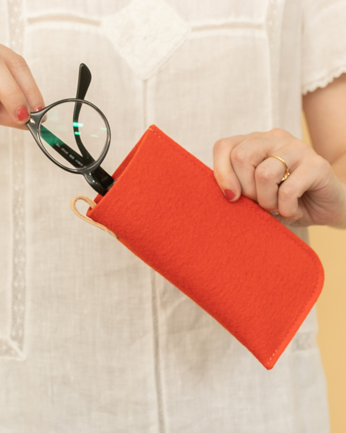 Wearing a white shirt and with red-painted nails, a person carefully places glasses into a graf lantz bright red felt case, an elegant sleeve designed for perfect eyewear protection, skillfully managing the case in one hand while holding the glasses in the other.