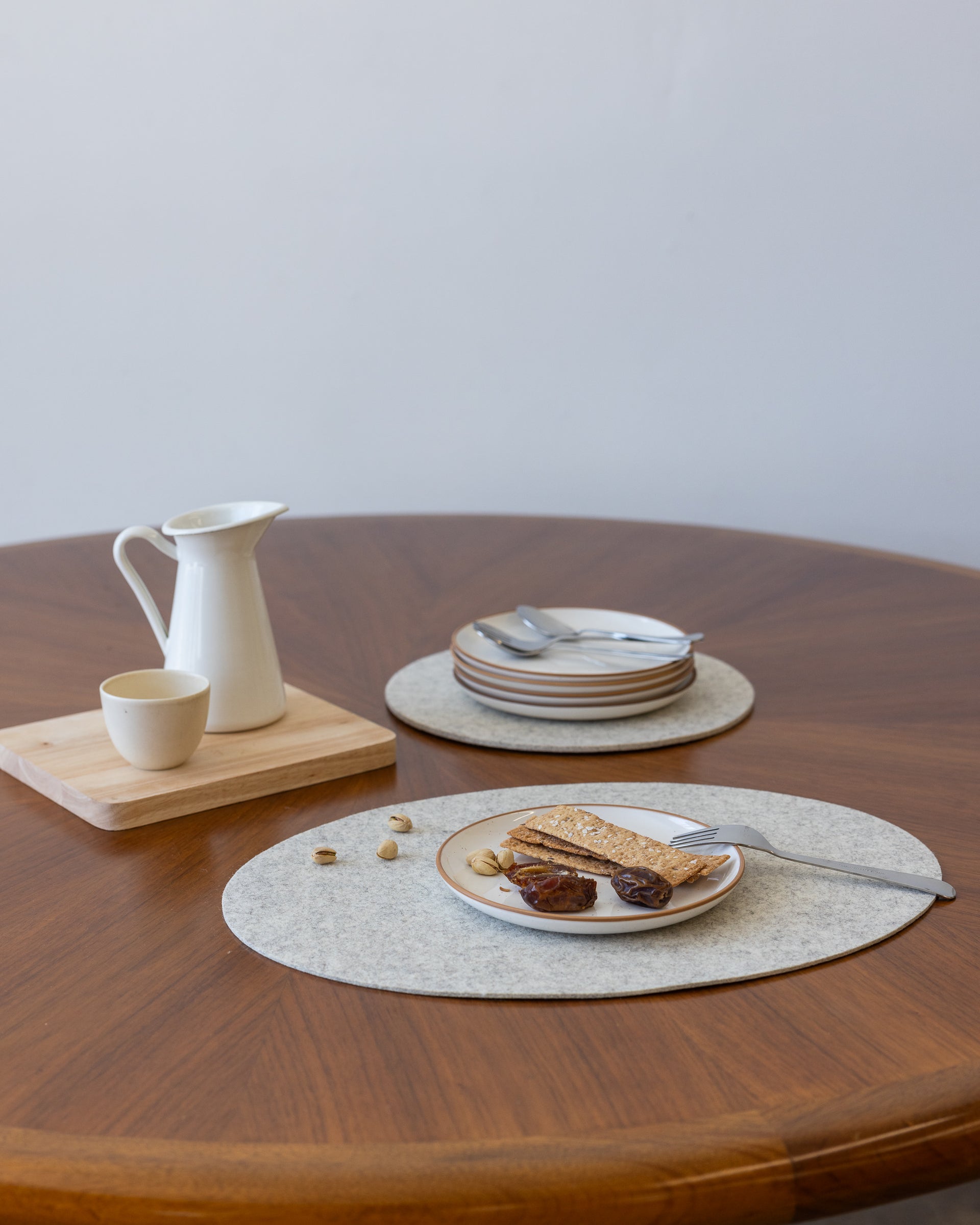 A round table features a white pitcher and cup on a wooden tray, with a nearby stack of plates and cutlery ready for use. Toast, figs, and nuts are artfully placed on a heat-resistant Merino Wool Placemat by graf lantz in the foreground.