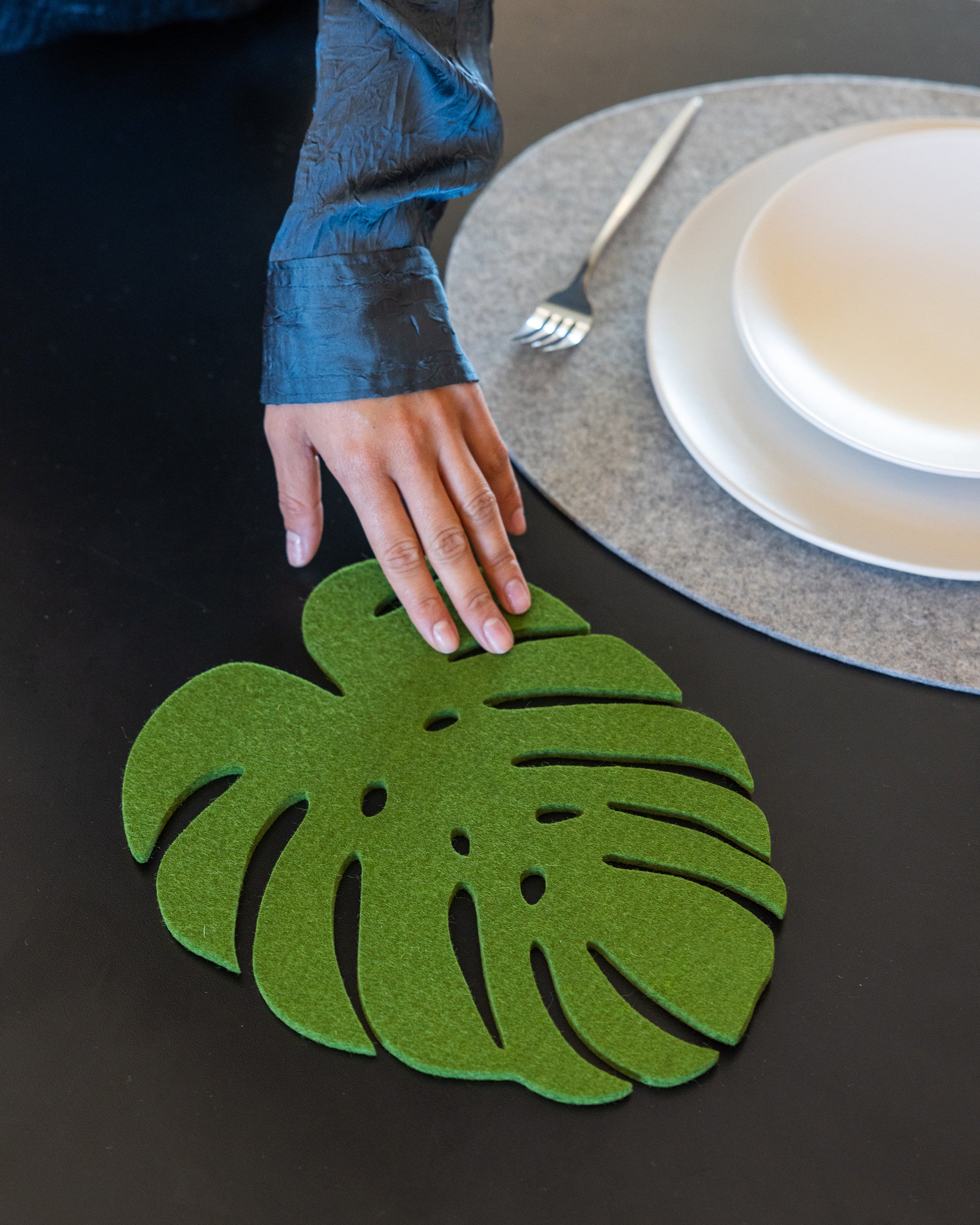 A hand in a dark blue long-sleeve shirt places a graf lantz sustainable green monstera-shaped felt placemat on a black table, while stacked white plates and a fork sit above on a gray circular mat.