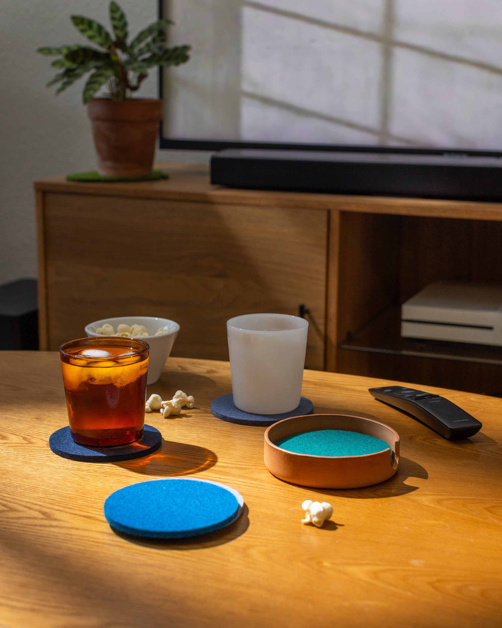 A cozy living room scene with iced tea and milk on coasters atop a graf lantz leather tray; popcorn scattered around and a visible remote. Sunlight brightens the area, while a potted plant perches on a nearby shelf.
