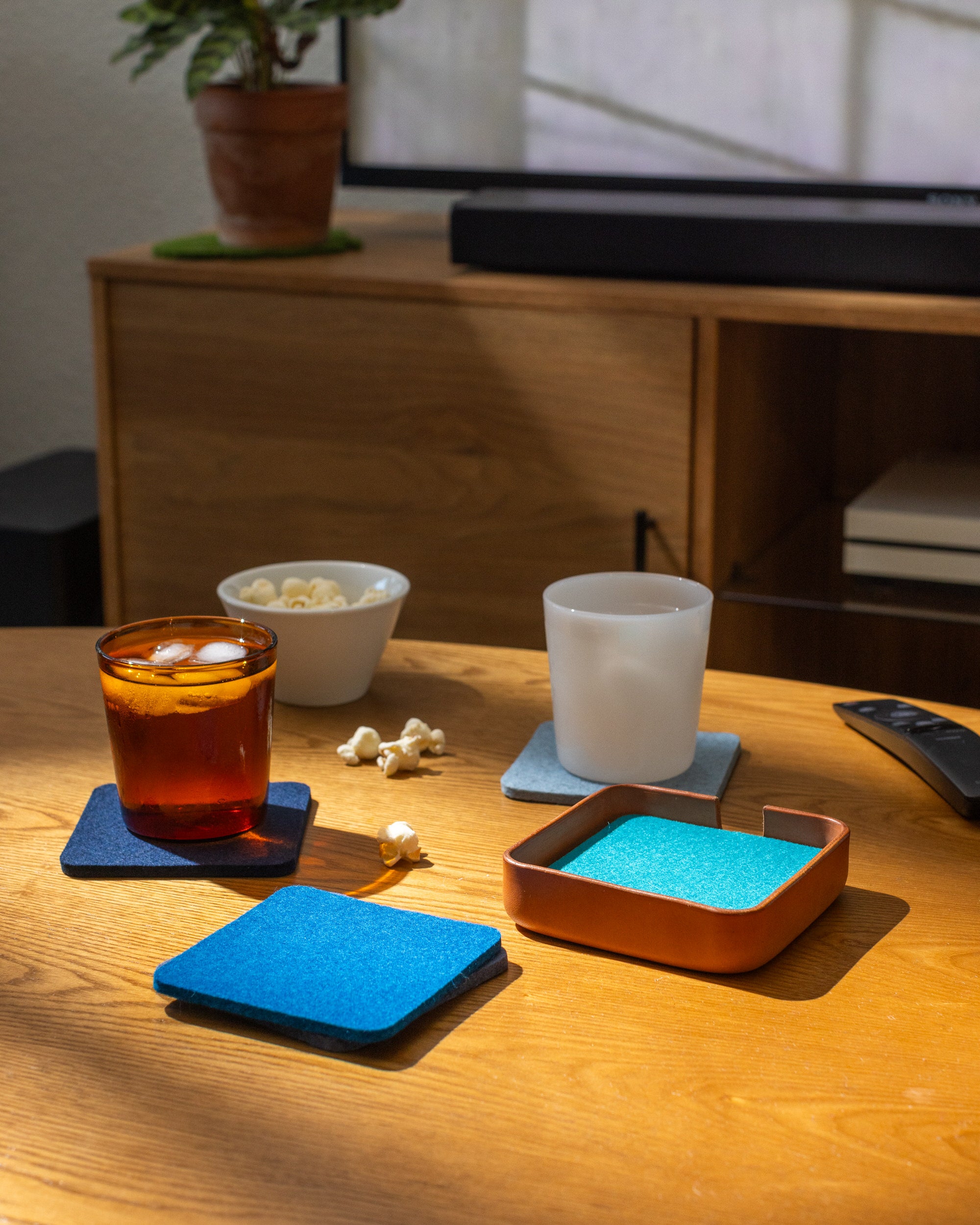 A cozy wooden table showcases a handcrafted glass of iced tea and a white mug placed on graf lantzs blue coasters. Nearby, a small bowl of popcorn complements the scene while sunlight filters through, casting shadows on the TV and bookshelf in the background.