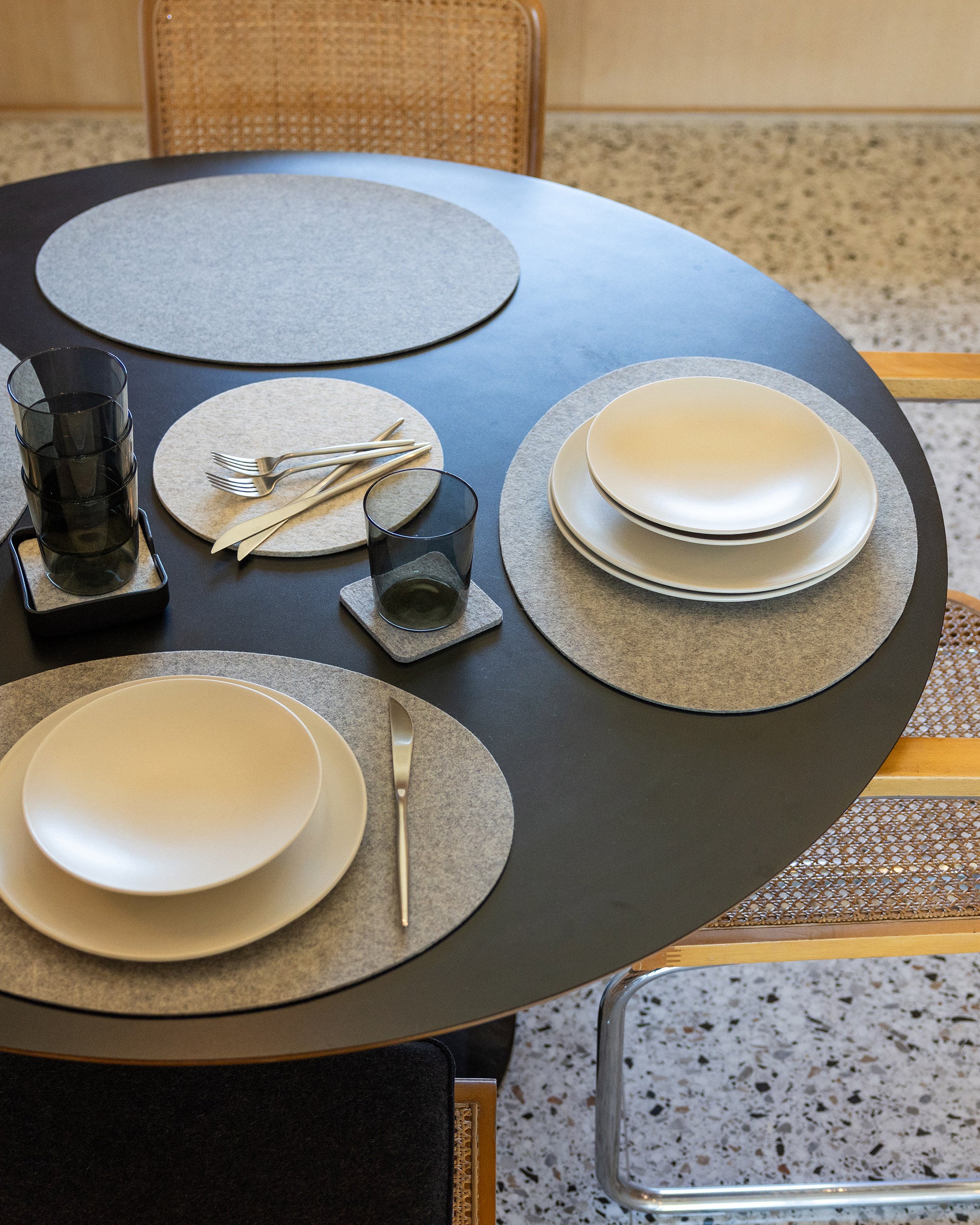 A round black dining table is adorned with graf lantz premium Merino wool placemats, white plates, and black glasses. Silverware rests on a beige napkin. Cane-backed chairs surround the table on a speckled floor.