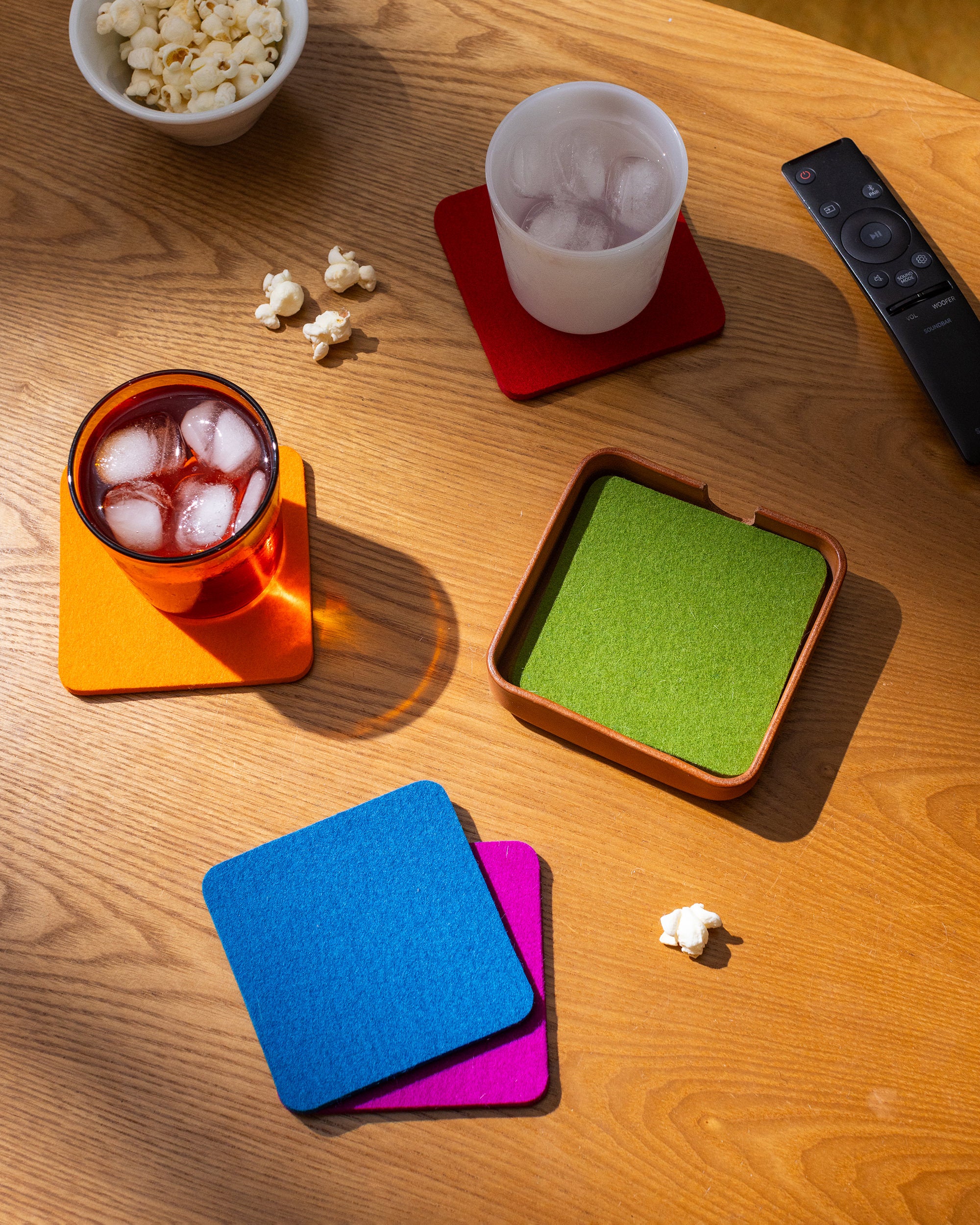 A wooden table showcases vibrant graf lantz Merino wool coasters: orange with a drink, red with a cup, and green in its holder. Nearby, a leather tray by Kobon holds blue and pink coasters. Popcorn and a TV remote complete the cozy scene.