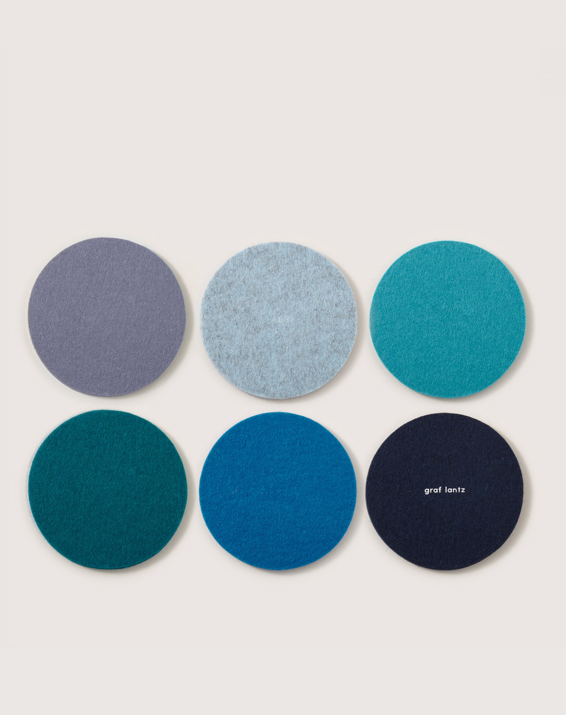 Six coasters, made from German merino wool, come in a sleek 3x2 layout by Graf Lantz. Each features unique blue and teal shades; one displays the Graf Lantz logo for stylish and elegant surface protection.