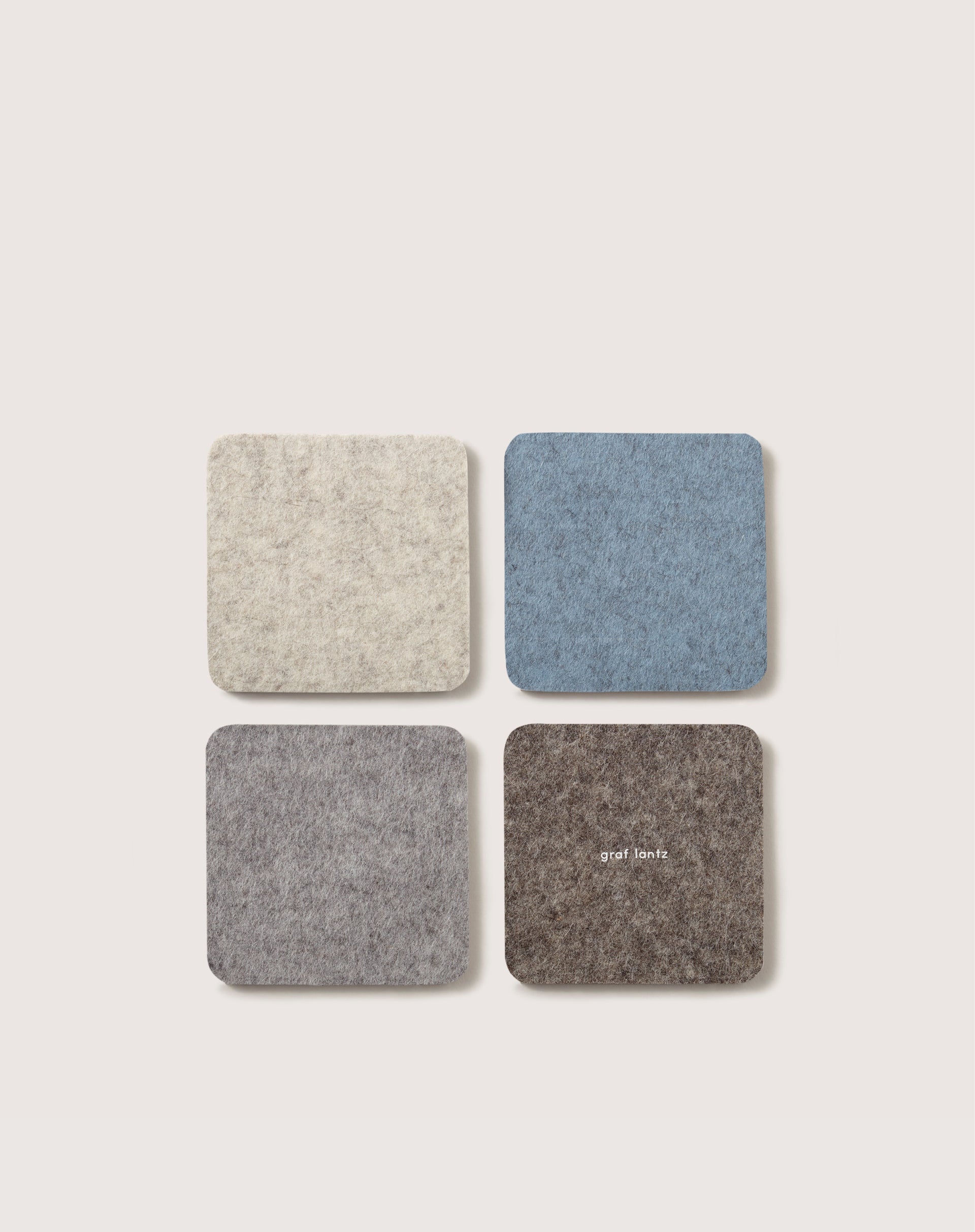 A set of four square felt coasters made from Merino wool, displayed in a grid on a light backdrop, featuring shades of light beige, blue, gray, and brown. The brown coaster bears the graf lantz brand name. Perfect for home décor.