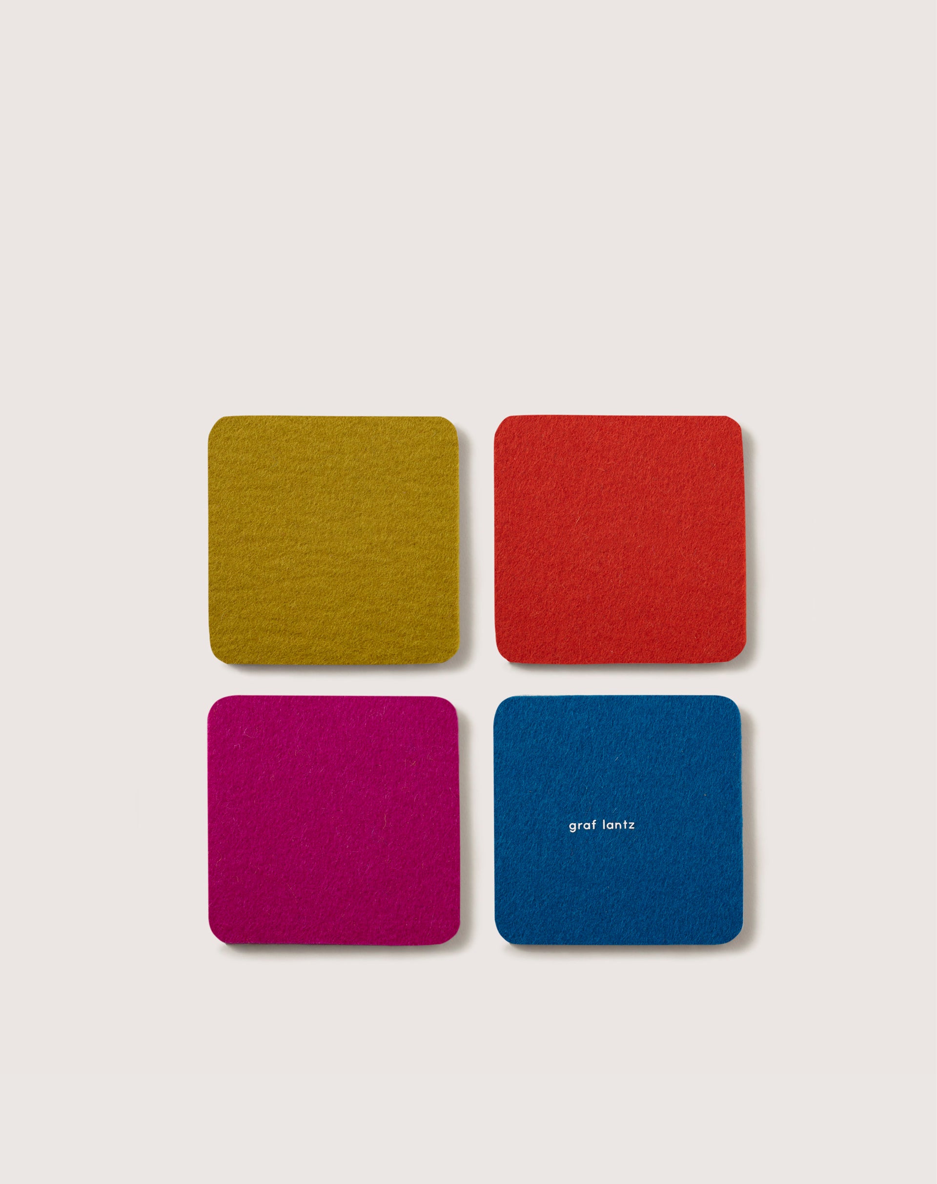 These four square felt coasters, crafted from premium Merino wool in vibrant hues of yellow, red, magenta, and blue (with graf lantz in white on the blue coaster), add a stylish touch to home décor or serve as essential beer mats. They are displayed neatly on a light backdrop.