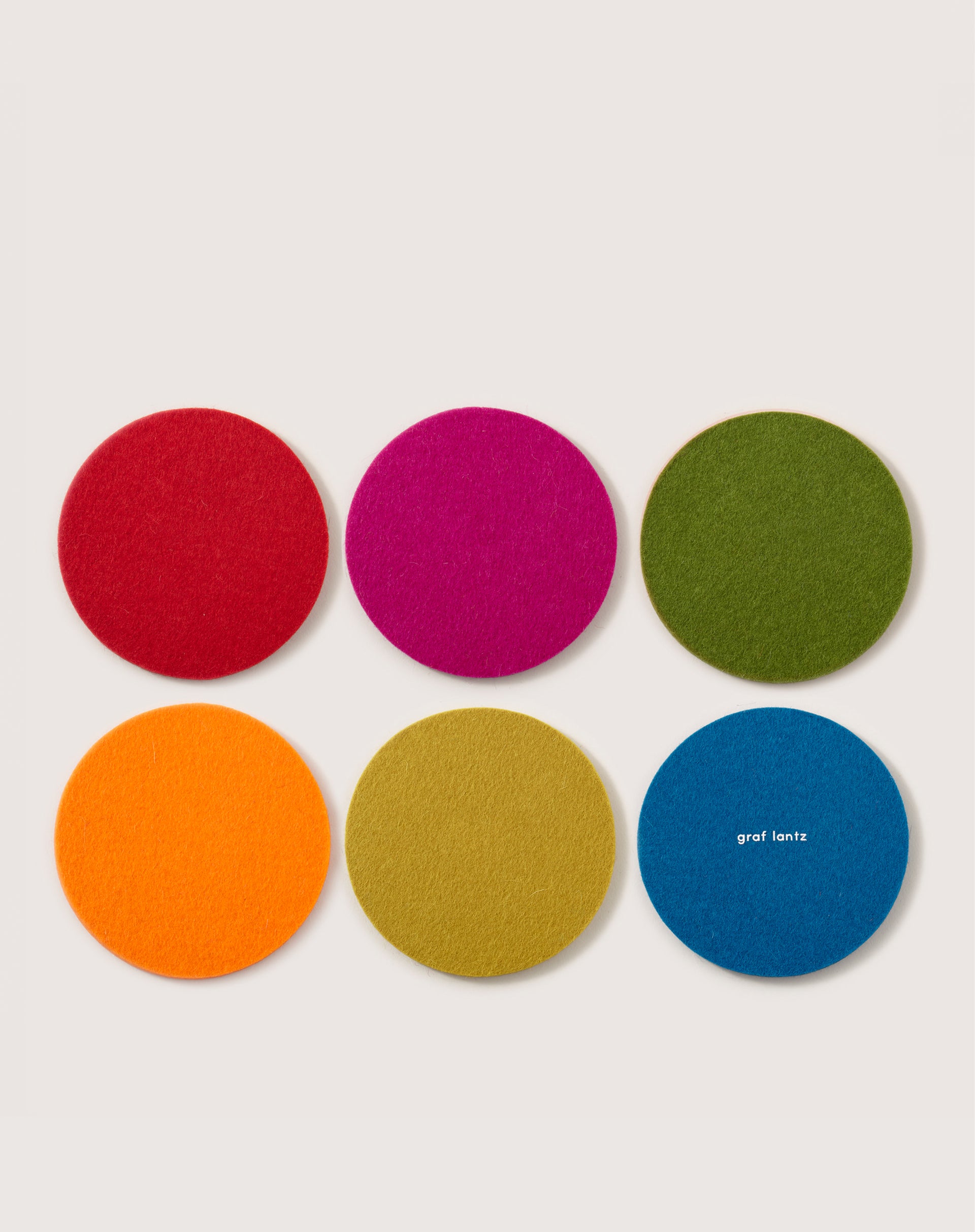 Six round coasters made from soft German Merino wool are arranged in two rows: red, pink, green on top; orange, yellow, blue on the bottom. The blue coaster features graf lantz against a light gray background.