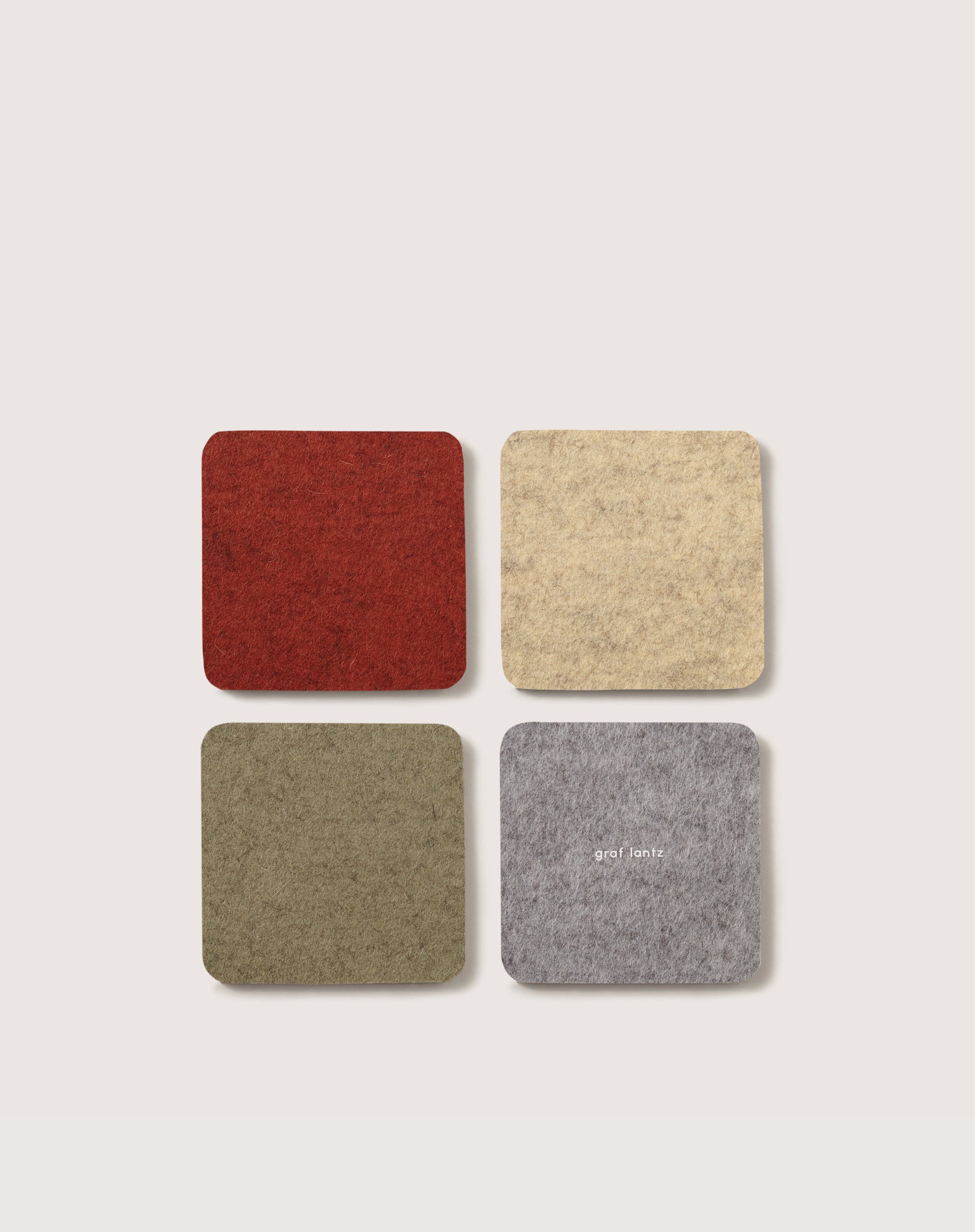 A set of four square graf lantz Merino wool coasters in red, beige, green, and gray arranged on a light background; the gray coaster features faint white text. They are perfect for safeguarding surfaces while enjoying your favorite Beer Felt moment.