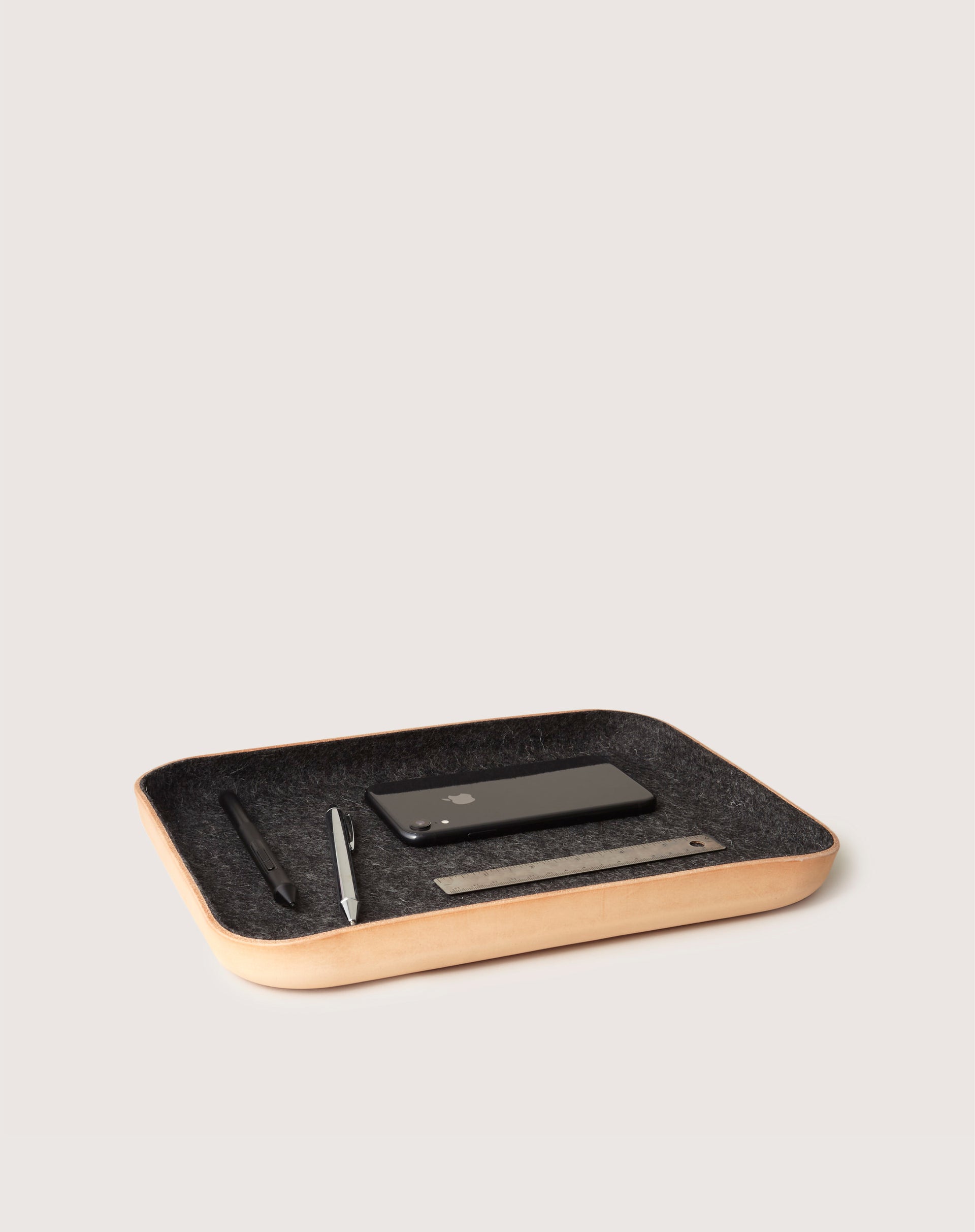 A graf lantz wooden catchall tray with Merino wool lining neatly organizes a smartphone, pen, metal ruler, and pencil against a plain white backdrop.