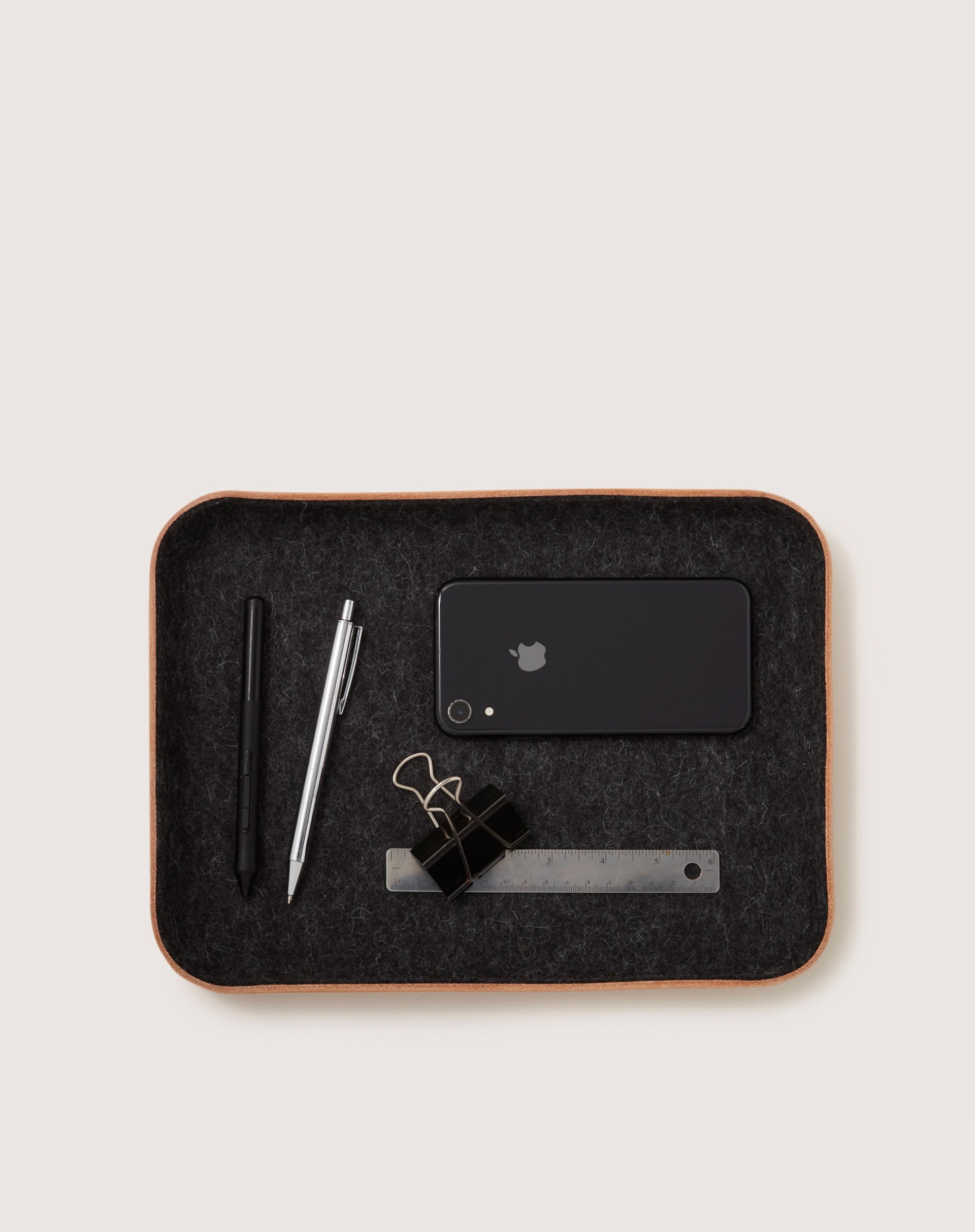 A graf lantz Merino wool catchall tray holds a black smartphone, two pens, two black binder clips, and a metal ruler against a neutral background.