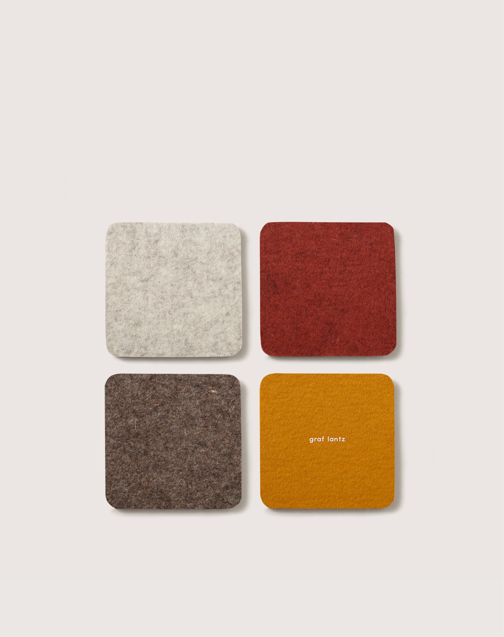 A set of four square coasters made from German Merino wool in light grey, red, brown, and mustard yellow is arranged in a grid pattern. The mustard yellow coaster subtly displays the brand name graf lantz.
