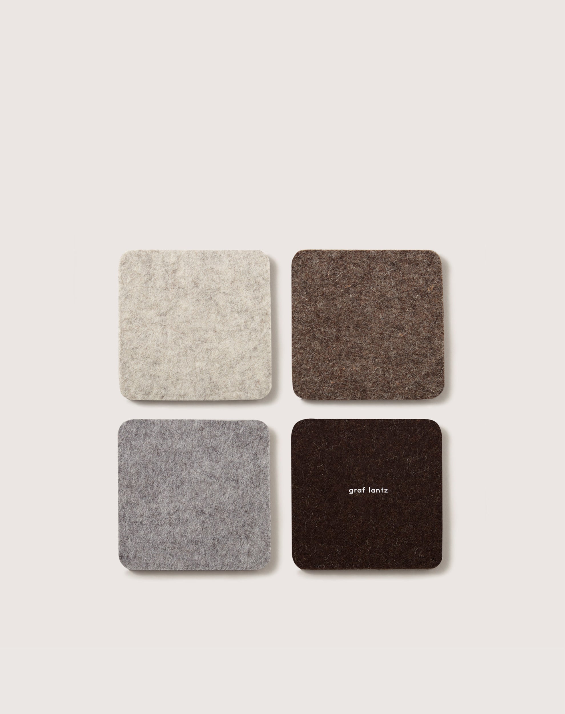 Four square felt coasters, crafted from premium German Merino wool in shades of gray and brown, are arranged on a light background. The coaster at the bottom right features the graf lantz brand name.