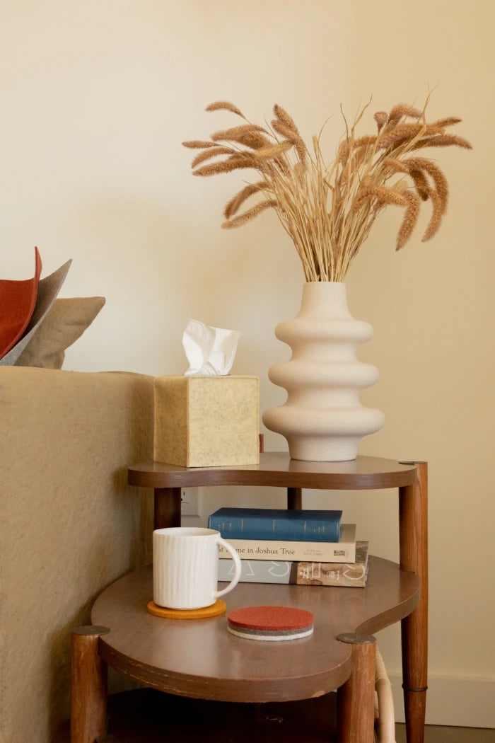 A cozy side table with two tiers features a wavy beige vase of dried grasses, books, a tan tissue box, and a white mug resting on a graf lantz Merino wool coaster. The scene beside the light couch exudes warmth and invites relaxation.