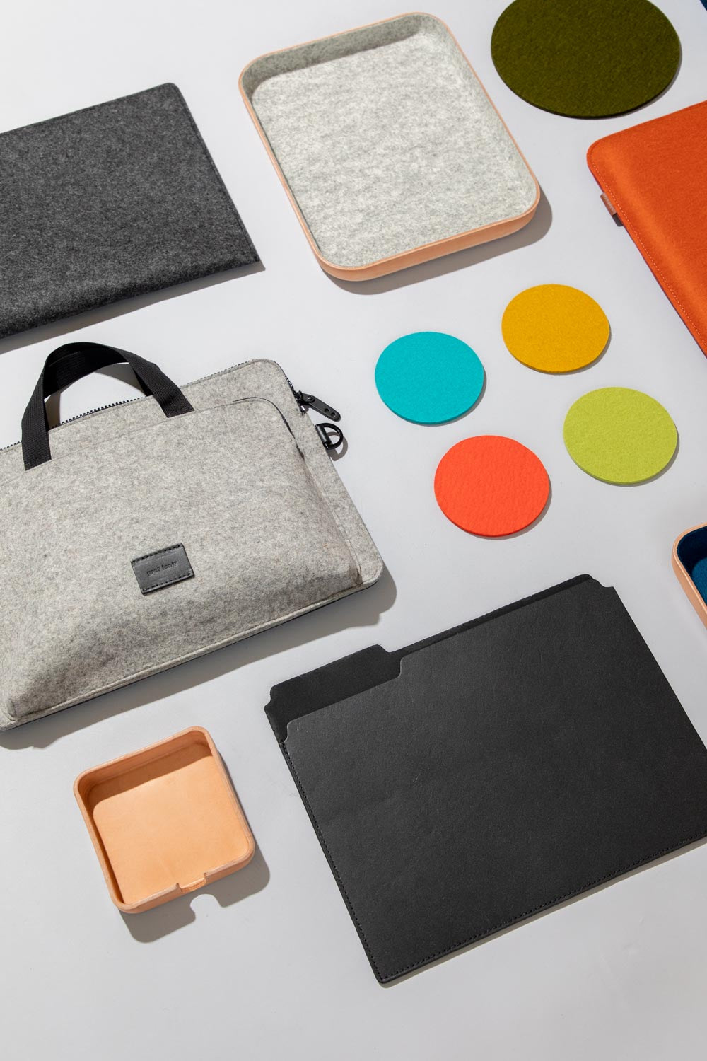A neatly arranged assortment of graf lantz office accessories on a light surface includes felt laptop sleeves, a leather file folder, colorful round coasters, a wooden tray, and a black document holder.